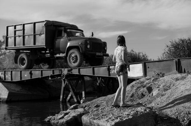 A girl watches and army truck pass by across a bridge over the river. She ahs just been swimming to cool of from summer.