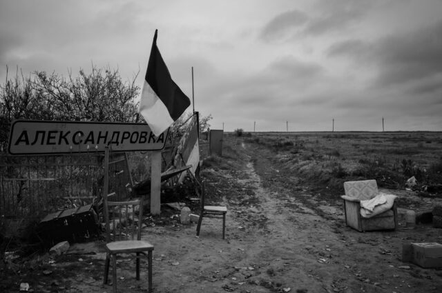 Landscape scene in Oleksandrvika with furniture outside, location sign and Ukrainian flag.