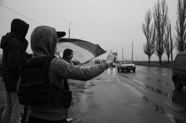 Youngsters waving the Ukrainian flag and making peace signs along the road to Kherson, it is raining and cars are passing by.
