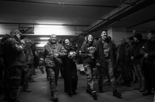 people and armed forces in uniform carrying flowers and smiling walking through a crowd at their wedding.