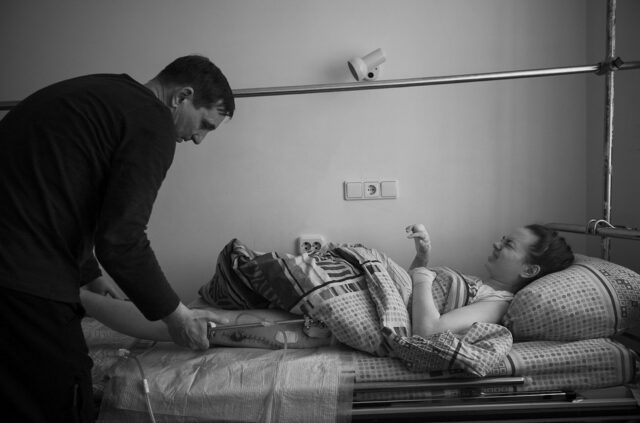 Nastya, lying in bed with pins, she is in pain, was evacuated from Chernihiv, two hours from Kyiv, on 22 March. During an airstrike she lost part of her leg and severely wounded the other. Her father tries to make her more comfortable, Kyiv, Ukraine - 25/04/2022.